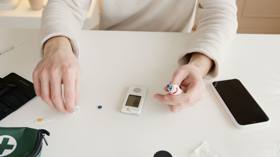 A person wearing a cream-colored long-sleeve shirt is seated at a white table, preparing to test soil or plant health in a garden. They are holding a small white device with a digital screen, likely a